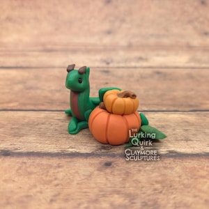 May include: A small green dragon figurine with brown horns and a brown belly stripe sitting next to two stacked orange pumpkins with green leaves. The text The Lurking Quirk & Claymore Sculptures is printed in white on the lower right side.
