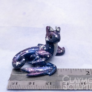 May include: A small, galaxy-themed dragon figurine with a blue, purple, and white paint scheme. The dragon is sitting on a ruler, measuring approximately 2 inches in length.