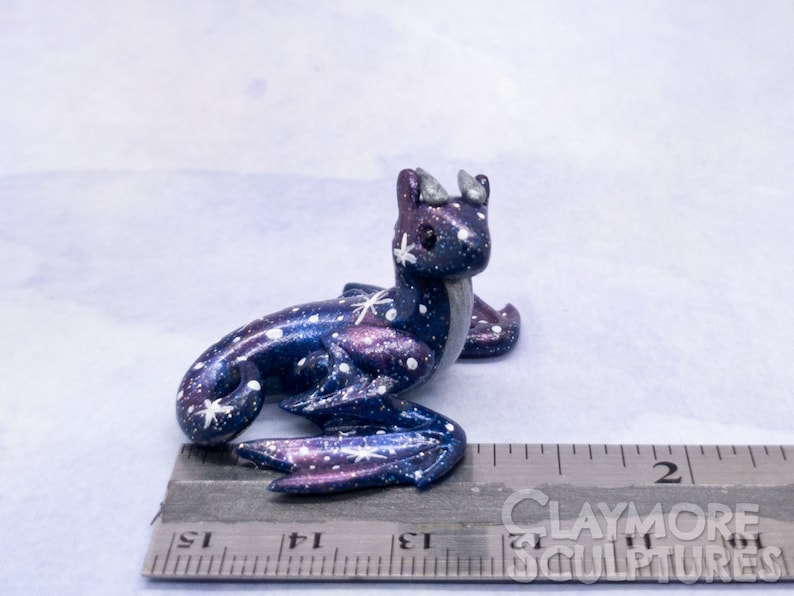 May include: A small, galaxy-themed dragon figurine with a blue, purple, and white paint scheme. The dragon is sitting on a ruler, measuring approximately 2 inches in length.