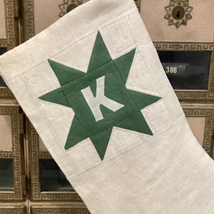 May include: A cream-colored Christmas stocking with a green starburst design and the letter "K" in the center. The stocking has a brown loop for hanging. The background features a vintage postal box.