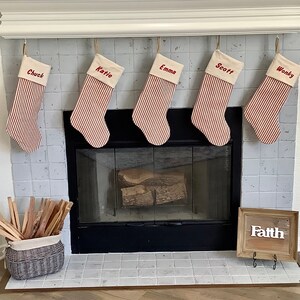 May include: Five personalized Christmas stockings with red and white stripes hanging above a fireplace. The stockings are labeled "Chuck", "Katie", "Emma", "Scott", and "Wonky". A wooden sign with the word "Faith" is on the mantle below the stockings.