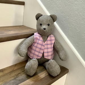 May include: A gray teddy bear wearing a pink and blue plaid vest. The bear is sitting on wooden stairs.