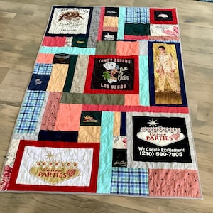 May include: A patchwork quilt with various fabric squares in shades of blue, pink, red, and beige. The quilt features logos and text from Tommy Bahama, Gerald's Casino Parties, and other brands. The quilt is rectangular and appears to be handmade.