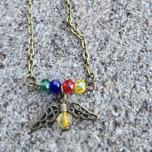 May include: A bronze necklace with a pendant of wings and a yellow bead. Above the pendant are four colourful beads: green, blue, red, and yellow. The necklace has a delicate chain and a clasp.