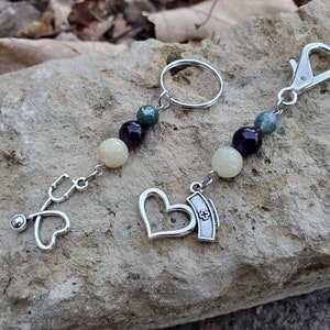 May include: A silver key chain with a stethoscope, heart, and nurse's cap charm. The chain is adorned with green, white, and purple beads.
