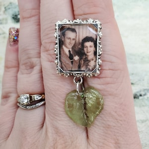 May include: A silver ring with a small picture frame charm featuring a black and white photo of a couple. A green leaf charm hangs from the frame.
