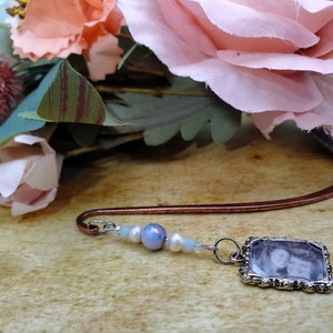 May include: A copper bookmark with a silver charm featuring a black and white photo of two people. The charm is surrounded by a decorative silver frame. The bookmark is adorned with blue, white, and pearl-colored beads.