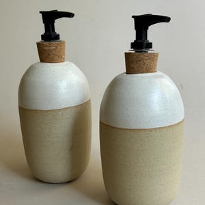 May include: Two ceramic soap dispensers with a rounded, two-tone design. The upper portion is a speckled white glaze, while the lower section is a natural, speckled beige clay. Each dispenser features a cork collar and a black plastic pump.
