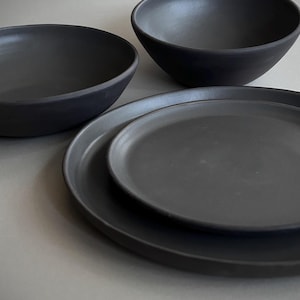 May include: A set of matte black dinnerware consisting of two circular plates of varying sizes and two deep bowls. The items feature a smooth, uniform dark finish with simple, rounded edges and a clean, functional design for table service.