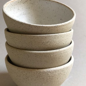 Pode incluir: Uma pilha de quatro pequenas tigelas de cerâmica salpicada. As tigelas são de cor bege claro com salpicos escuros e uma textura ligeiramente áspera. As tigelas estão empilhadas umas sobre as outras, com a tigela superior ligeiramente inclinada.