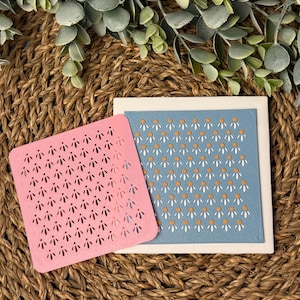May include: Two square stencils, one pink and one blue, with a daisy pattern. The pink stencil is cut out, while the blue stencil has a painted daisy pattern.