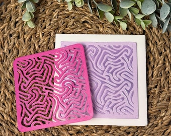 May include: Two square silicone molds with a raised, abstract, maze-like design. The mold on the left is pink and the mold on the right is purple.