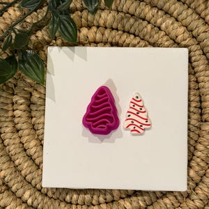 May include: A fuchsia plastic Christmas tree-shaped cookie cutter next to a small, decorated Christmas tree. The tree is white with red stripes and a green top. The items are on a white square.