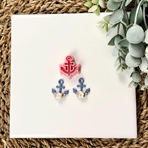 May include: Three nautical-themed items: a pink anchor-shaped cookie cutter and two blue anchor earrings with floral accents. The items are arranged on a white square surface, with a woven background and greenery.