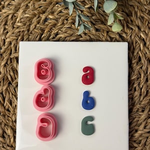 May include: Pink letter-shaped cookie cutters and colourful clay letters. The cutters are in the shape of the letter "B". The clay letters are red "a", blue "b", and green "c". The items are on a white square surface.