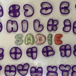 May include: A collection of purple plastic cookie cutters in various letter shapes arranged in rows, surrounding five three-dimensional ceramic letters that spell out the name SADIE, each featuring distinct colorful patterns and designs.