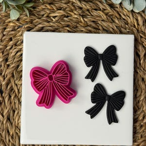 May include: A pink bow-shaped cookie cutter next to two black bow-shaped decorations. The cookie cutter is made of plastic and has a detailed design. The black bows appear to be made of a rubbery material.