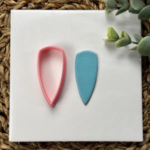 May include: A pink plastic cookie cutter and a light blue polymer clay leaf shape on a white background.