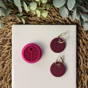 May include: A fuchsia clay cutter and two burgundy earrings with gold hoops are shown on a white surface. The earrings and cutter have a geometric design of parallel lines. The background includes greenery and a woven texture.