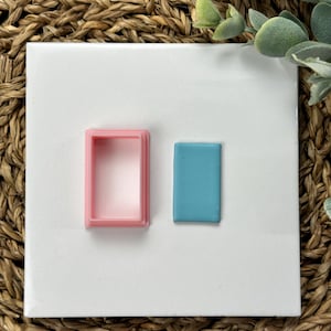 May include: A pink rectangular cookie cutter next to a small, rectangular piece of blue clay. The cookie cutter is empty, showing its shape. The clay is a solid color and fits inside the cutter. The items are on a white surface.