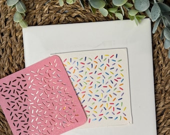 May include: Two square cards with a pattern of colorful sprinkles. The card on the left is pink and the card on the right is white.