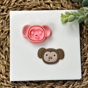 May include: A pink monkey-shaped cookie cutter and a brown monkey-shaped embellishment are displayed on a white square. The cookie cutter is made of pink plastic, and the embellishment is brown with a tan face and pink nose.