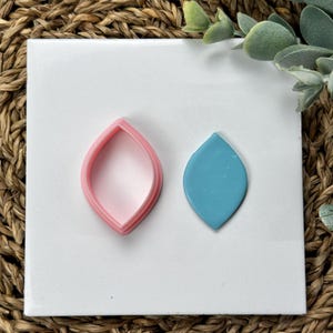 May include: A pink leaf-shaped cookie cutter and a teal leaf-shaped polymer clay cutout on a white background.
