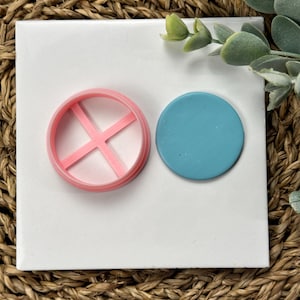 May include: A pink plastic cookie cutter with a cross-shaped design and a solid blue circle of polymer clay.