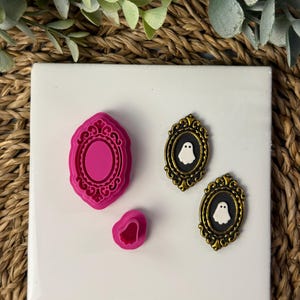 May include: A pink, ornate, oval-shaped mold and a smaller, matching cutter sit next to two antique-style, black and gold framed ghost pendants. The pendants feature a white ghost on a black background. The items are on a white tile.