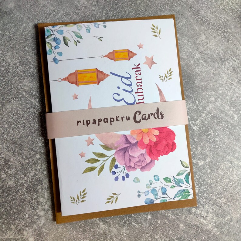 May include: A white card with a crescent moon, stars, and lanterns. The card has a floral design with pink, red, and purple flowers. The text "Eid Mubarak" is written in blue and the text "ripapaperu Cards" is written in brown.