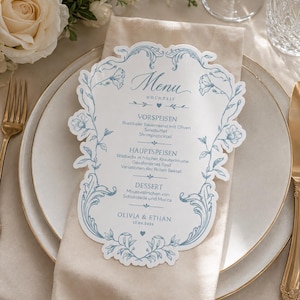 May include: A die-cut wedding menu card with blue floral scrollwork borders on a white background. The text reads Menu, Hochzeit, Vorspeisen, Hauptspeisen, Dessert, and the names Olivia & Ethan with the date 12.08.2026. The card is placed on a beige napkin.