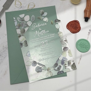 May include: A clear acrylic wedding invitation with a green and white floral design. The invitation reads "Sabrina & Matthea" and includes the date "May 27, 2023" and the time "13:00 Uhr".