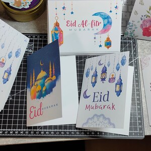 May include: Assortment of greeting cards with watercolor illustrations of crescent moons, lanterns, and floral designs. The cards feature the text "Eid Mubarak" and "Eid Al-Fitr Mubarak".