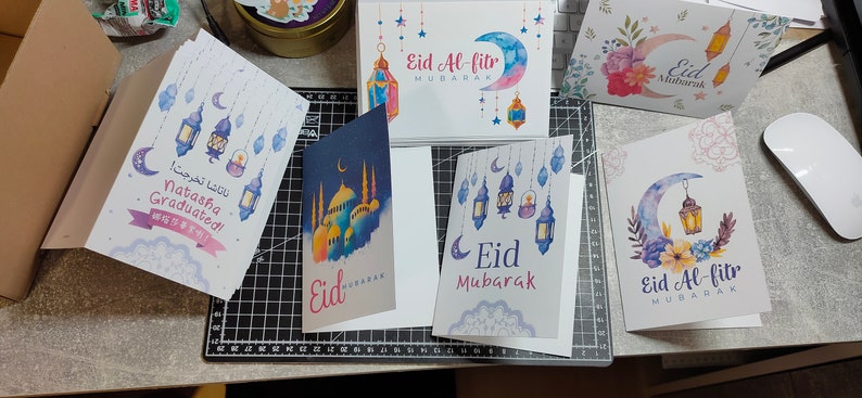 May include: Assortment of greeting cards with watercolor illustrations of crescent moons, lanterns, and floral designs. The cards feature the text "Eid Mubarak" and "Eid Al-Fitr Mubarak".