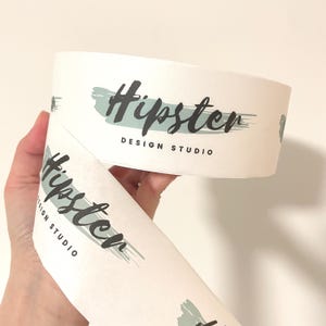Peut inclure: Rouleau de papier blanc avec l'inscription "Hipster DESIGN STUDIO" en écriture noire et un motif de coup de pinceau turquoise. Le papier est tenu dans une main.