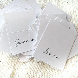 May include: A stack of white place cards with the names "Juana" and "Laura" written in black calligraphy.