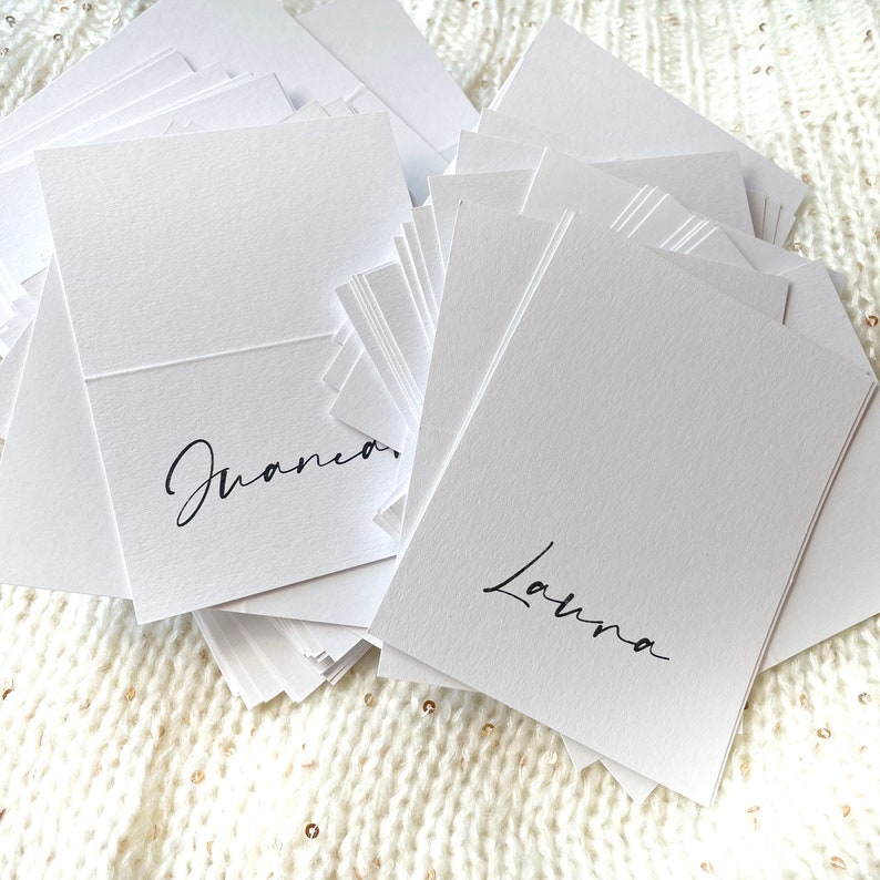 May include: A stack of white place cards with the names "Juana" and "Laura" written in black calligraphy.