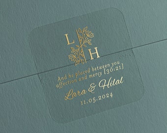 以下が含まれることがあります： 金色の文字と花の模様が入った透明な正方形のステッカー。ステッカーにはイニシャル「L」と「H」、名前「Lara & Hilal」、日付「11.05.2024」、引用句が記載されています。ステッカーはティール色の背景に配置されています。
