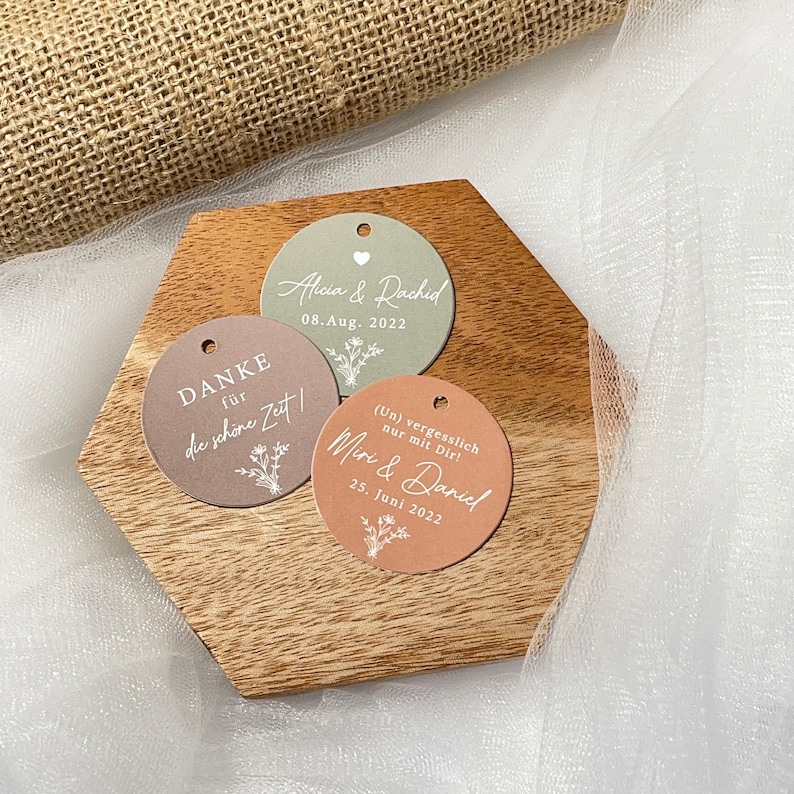 Puede incluir: Tres etiquetas de regalo redondas de papel con texto blanco y dise&ntilde;os florales. La etiqueta superior es verde y dice "Alicia & Rachid 08. Aug. 2022". La etiqueta del medio es marr&oacute;n y dice "DANKE f&uuml;r die sch&ouml;ne Zeit". La etiqueta inferior es naranja y dice "(Un) vergesslich nur mit Dir! Miri & Daniel 25. Juni 2022".