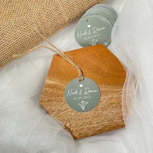 Puede incluir: Un conjunto de etiquetas redondas de color gris plateado para favores de boda con texto blanco. Las etiquetas muestran los nombres "Nicole & Roman" y la fecha "10.09.2022". Tambi&eacute;n se incluye un peque&ntilde;o coraz&oacute;n blanco y un dise&ntilde;o floral.