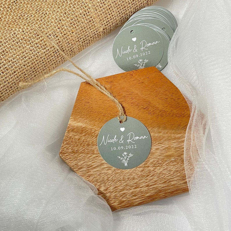 Puede incluir: Un conjunto de etiquetas redondas de color gris plateado para favores de boda con texto blanco. Las etiquetas muestran los nombres "Nicole & Roman" y la fecha "10.09.2022". Tambi&eacute;n se incluye un peque&ntilde;o coraz&oacute;n blanco y un dise&ntilde;o floral.