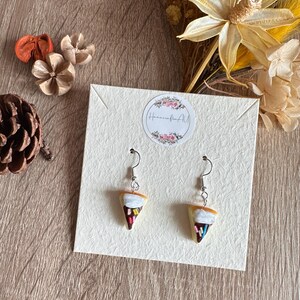 Könnte beinhalten: Ein Paar handgefertigte Eiscreme-Kegel-Ohrringe. Jeder Ohrring hat einen braunen Kegel, weißes Eis und bunte Streusel. Die Ohrringe werden auf einer cremefarbenen Karte mit einem floralen Logo präsentiert.