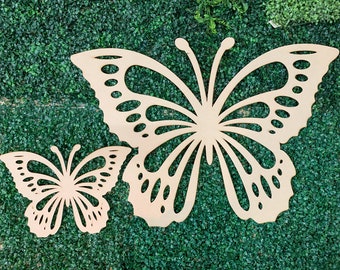 May include: Two wooden butterfly cutouts, one large and one small, are displayed against a green artificial grass background. The butterflies have intricate wing designs and are made of light brown wood.