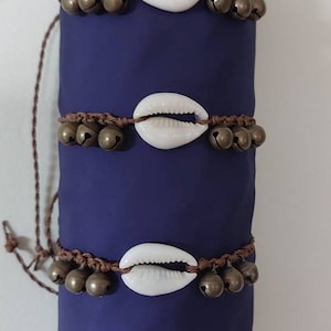 May include: Five brown cord bracelets with white cowrie shells and gold-toned bells. The bracelets are stacked on a blue background.