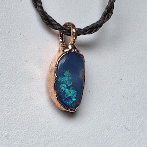 May include: A pendant featuring a dark blue stone with iridescent green and blue flecks, encased in a textured copper setting. The pendant is suspended from a black braided cord, creating a contrast between the metallic copper and the dark stone.