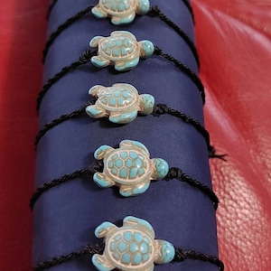 Könnte beinhalten: Ein Set mit sechs türkisfarbenen Schildkröten-Anhänger-Armbändern mit schwarzen Schnüren. Die Schildkröten sind aus glattem, poliertem Stein gefertigt und haben ein detailliertes Design.