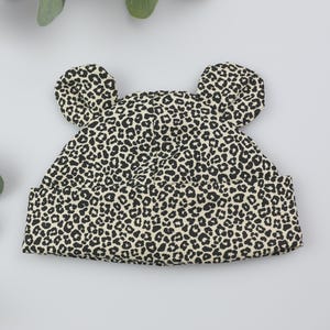 Peut inclure: Un bonnet pour bébé avec un motif léopard noir et beige. Le chapeau a deux petites formes arrondies en forme d'oreilles sur le dessus. Le chapeau est en tissu doux et est conçu pour s'adapter à la tête d'un bébé.