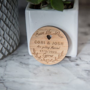 May include: A wooden save the date card with the text "Save the Date" and "Cori & Josh Are getting Married 17/5/2023 Cyprus" engraved on it.