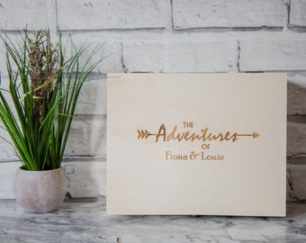 May include: A white wooden box with the text "The Adventures of Fiona & Louie" engraved on the front. The text is in a cursive font with a small arrow on either side.