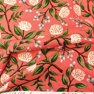 May include: Coral pink fabric with a white floral pattern. The fabric features a repeating design of white flowers with green leaves and stems. The fabric has a white label with the text "Wildwood Collection" printed on it.
